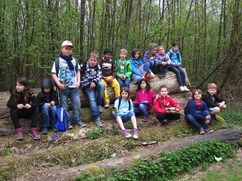 Das Foto zeigt die Kinder und vor einem großen Baumstamm sitzend, einige haben gerade ihre GPS Empfänger in der Hand und orientieren sich.